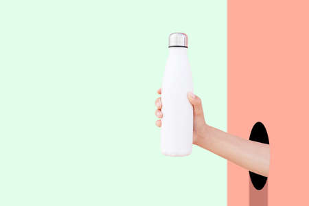 Close-up of hand through hole holding eco reusable steel thermo water bottle of white against red and green backgrounds with copy space.の写真素材