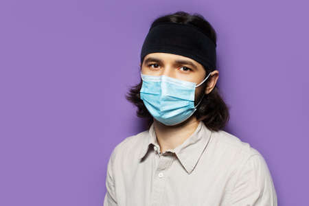 Studio portrait of young brunette man, with long hair, wearing protective medical mask. On the background of purple wall.の写真素材