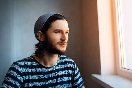 Portrait of young man looking outside through window, wearing beanie hat.