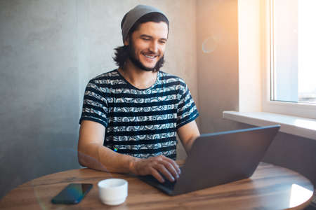 Indoor portrait of young confident smiling man, with grey beanie hat and striped T-shirt, typing on the laptop.の写真素材