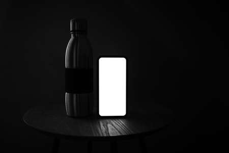 Dark photo in black and white of smartphone with mockup and steel reusable thermo bottle on wooden table against black background.の写真素材