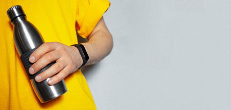 Close-up of male hand wearing smartwatch and holding steel reusable bottle in yellow shirt on background of grey wall with copy space. Focus on smartwatch.の写真素材