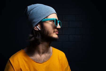 Studio side portrait of young confident man with wireless earphones wearing yellow t-shirt, blue sunglasses and grey hat on black background.の写真素材