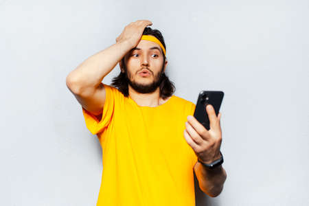 Portrait of surprised young man looking in smartphone wearing yellow shirt on background of textured white wall.の写真素材
