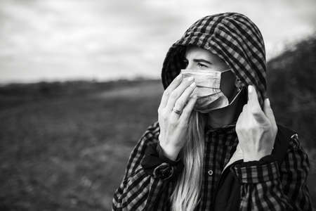 Black and white portrait of young hooded girl putting on medical face mask against coronavirus and...