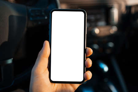 Close-up of male hand holding smartphone with white mockup on screen against blurred background of car dashboard.の写真素材