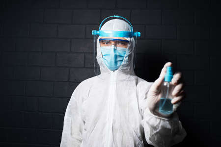 Portrait of doctor wearing PPE suit against coronavirus and covid-19, holding sanitizer bottle spray on background of black brick wall.の写真素材