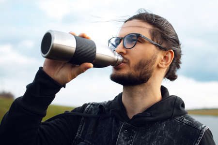 Portrait of young man drinking water from reusable metal bottle on background of blurred cloudy sky.の素材 [FY310170309969]