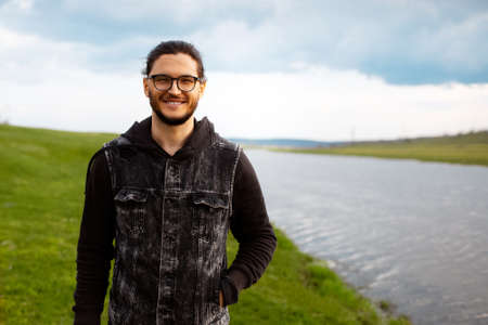 Outdoor portrait of young smiling man near river on background of blurred green field and cloudy sky.の写真素材