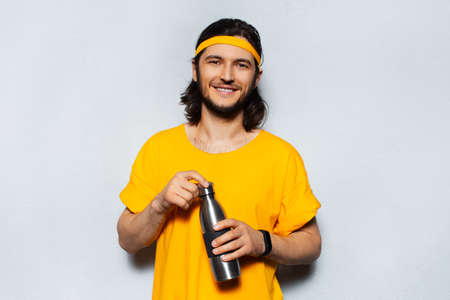 Portrait of young smiling man holding reusable steel thermo water bottle on background of grey wall....