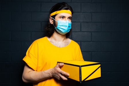 Portrait of young delivery man holding a yellow box, wearing medical mask and orange shirt on background of black brick wall.の写真素材