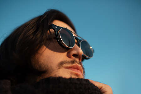 Close-up portrait of young guy with long hair wearing hipster round sunglasses and scarf. Blue sky...