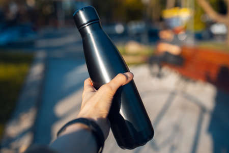 Close-up of female hand holding steel thermo water bottle of black. Metal eco reusable bottles concept; zero waste. Background of blurred street.の写真素材