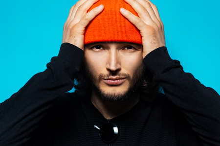 Close-up portrait of young confident man, holding hands on head. Wearing orange hat and black sweater. Studio photo on background of blue color.の写真素材