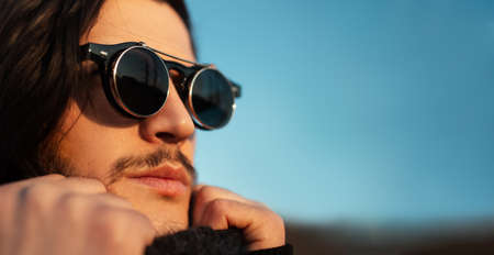 Close-up portrait of young guy with long hair wearing hipster round sunglasses and scarf. Blue sky on background. Panoramic banner view with copy space.の写真素材