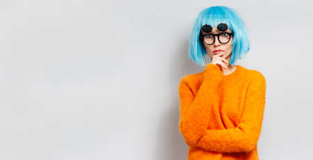 Studio portrait of young thoughtful girl on background of white background. Wearing round sunglasses, blue wig and orange sweater. Panoramic banner view with copy space.の写真素材