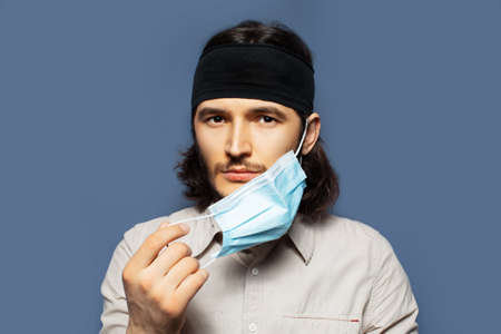 Studio portrait of young guy taking off medical face mask on background of blue color. End of...