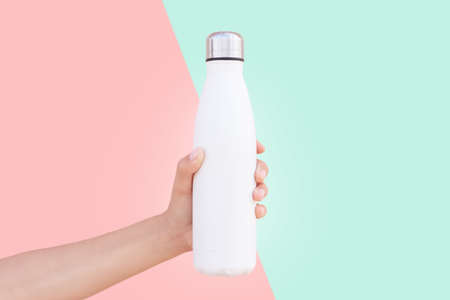 Close-up of female hand holding white reusable steel thermo water bottle isolated on two background of red and green colors.の写真素材