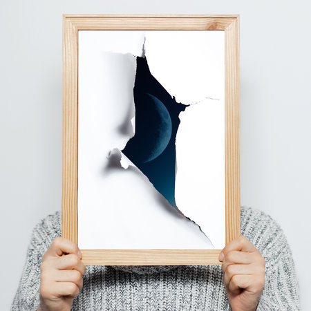 Close-up portrait of young man holding a wooden frame with view of moon through torn hole. Background of grey color.の写真素材