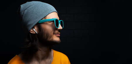 Close-up portrait of young smiling man with wireless earphone in ear on black background; wearing blue sunglasses. Panoramic banner view with copy space.の写真素材