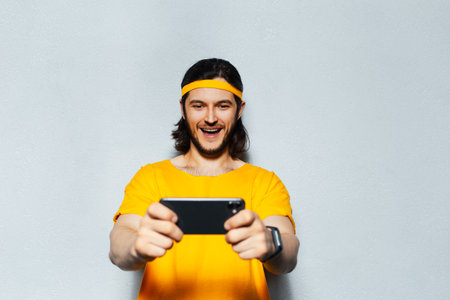 Studio portrait of young happy man in yellow with long hair, playing game on smartphone on grey background.の写真素材