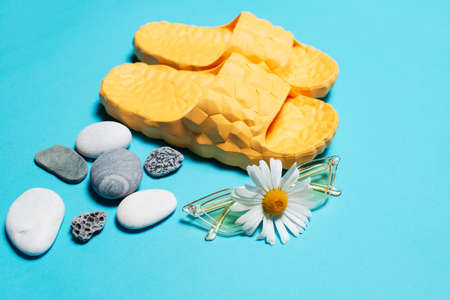 Close-up of chamomile flower on yellow sunglasses over blue background and slippers near pebbles.
