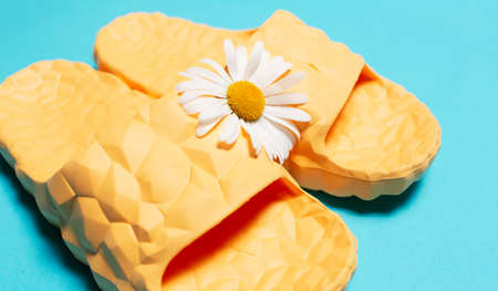 Close-up of yellow modern slippers and white chamomile flower on blue background.
