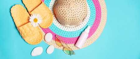 Top view of beach set accessories, woman summer hat, yellow slippers, sunglasses and cream tube decorated with chamomile flower and smooth white pebbles on blue background. Panoramic banner view.の写真素材