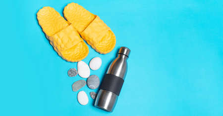 Top view of reusable steel thermo water bottle near yellow slippers and stones on blue background.の写真素材