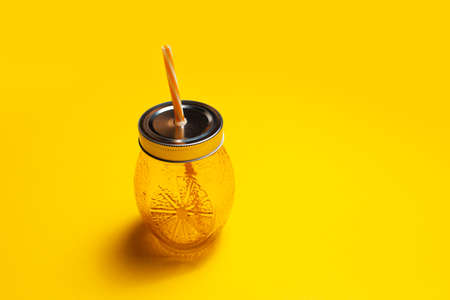 Close-up of juice glass mug of orange color, with straw and steel cap, on yellow background.