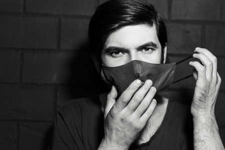 Young man taking off medical mask from face on background of black brick wall.の写真素材