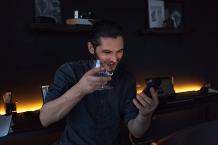 Happy guy talking online by smartphone. Cheers with cognac glass in hand.