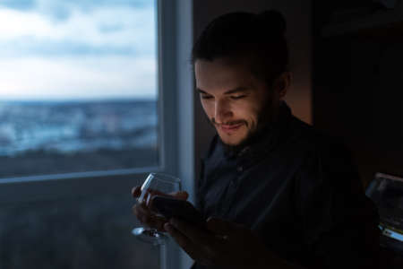 Portrait of young man looking in smartphone with cognac glass in hand.