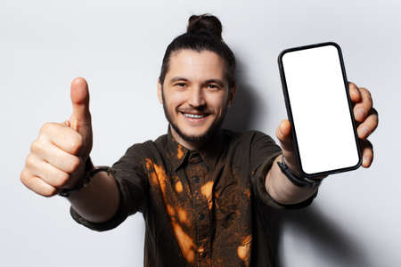 Young happy man with hair bun, showing thumb up and smartphone with mockup.