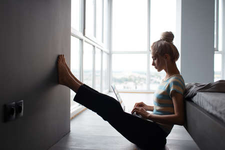 Blonde girl with hair bun working home, typing on laptop, sitting on the floor.の写真素材