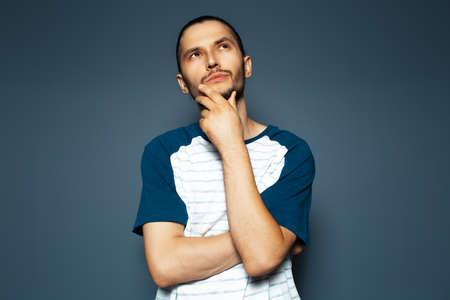 Studio portrait of young thoughtful man looking up with hand under chin on blue background.の写真素材