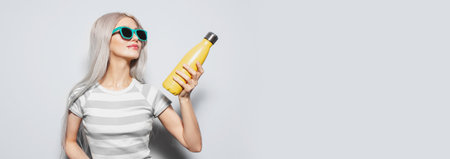 Studio portrait of young pretty blonde girl in striped shirt holding metal thermo water bottle of yellow color on white background with copy space, wearing blue sunglasses. Panoramic banner view.の写真素材