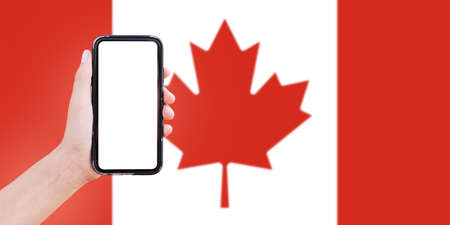 Close-up of male hand holding smartphone with blank on screen, on background of blurred flag of Canada.の写真素材