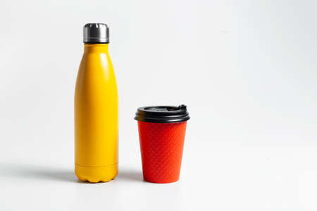 Close-up of metal thermo water bottle of yellow near paper cup for coffee takeaway of red color, on white studio background.の写真素材