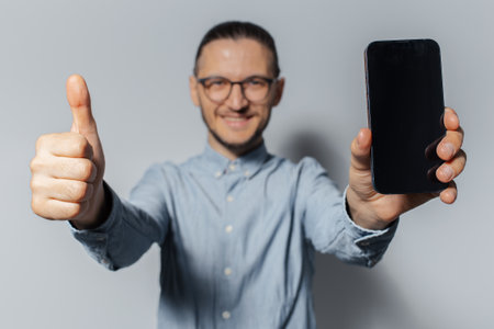 Studio portrait of young smiling man, showing thumb up and smartphone close to the camera, wearing eyeglasses and blue t-shirt on white background.の写真素材