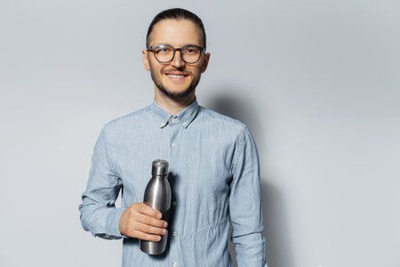 Studio portrait of young smiling guy holding metal bottle in hand on white background, wearing eyeglasses and blue shirt.の写真素材