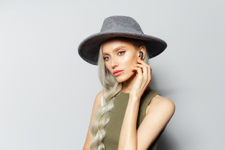 Studio portrait of cute blonde girl touching wireless earphones while listening to music, on white background. Wearing grey hat.の写真素材