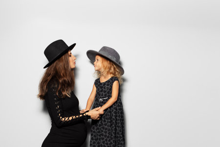 Studio portrait of pregnant smiling woman and child girl looking each ever, wearing hats on white background. Mother and daughter in black.の写真素材