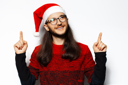Studio portrait of young smiling man in red sweater, wearing Santa Claus hat and eyeglasses, pointing fingers up, looking up. Isolated on white background.の写真素材