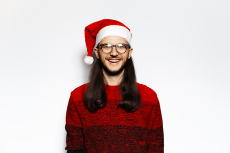 Studio portrait of young happy man in red sweater, wearing Santa Claus hat and eyeglasses. Isolated on white background.の写真素材