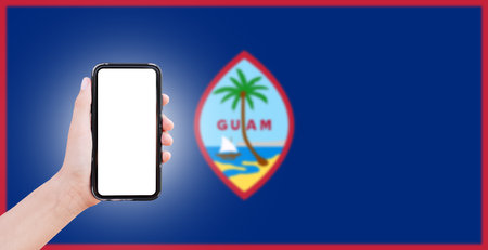 Male hand holding smartphone with blank on screen, on background of blurred flag of Guam. Close-up view.の写真素材