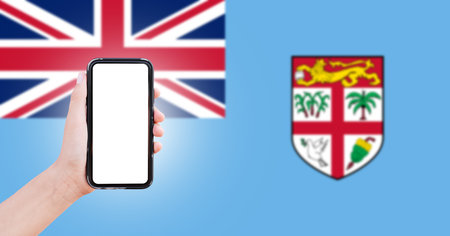 Male hand holding smartphone with blank on screen, on background of blurred flag of Fiji. Close-up view.の写真素材