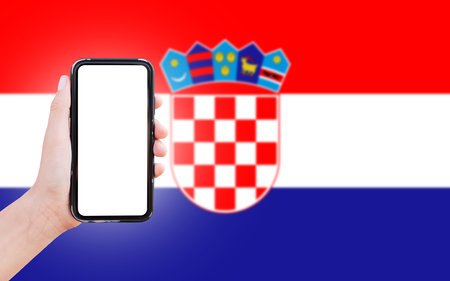 Male hand holding smartphone with blank on screen, on background of blurred flag of Croatia. Close-up view.の写真素材