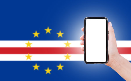 Male hand holding smartphone with blank on screen, on background of blurred flag of Cape Verde....