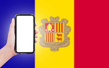 Male hand holding smartphone with blank on screen, on background of blurred flag of Andorra. Close-up view.の写真素材